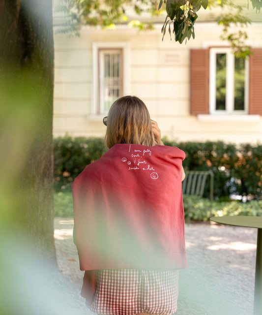 A person with shoulder-length hair stands outdoors wearing a limited edition red PRETTY COOL Jumper by WALD Berlin, featuring white text. A residential building and greenery in the background add a touch of tranquility to the scene.
