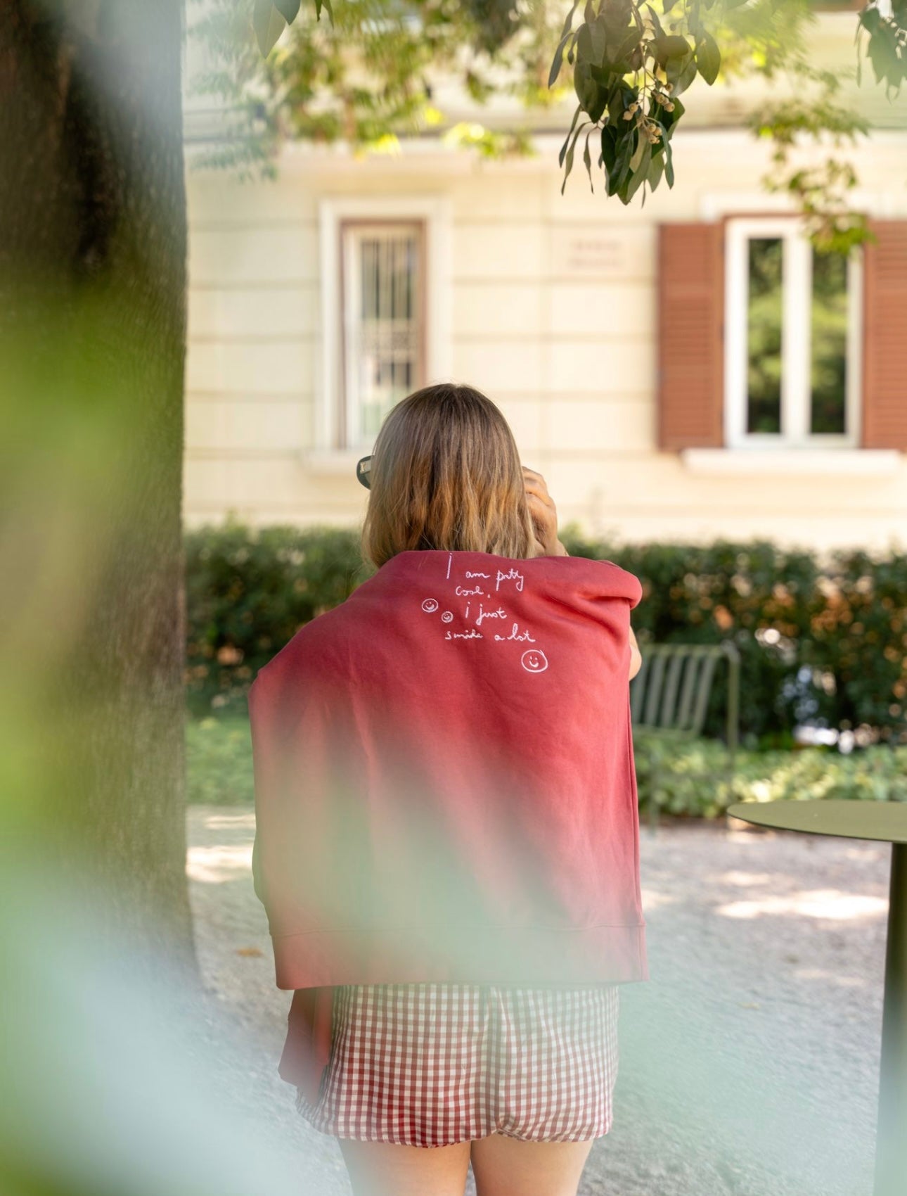 A person with shoulder-length hair stands outdoors wearing a limited edition red PRETTY COOL Jumper by WALD Berlin, featuring white text. A residential building and greenery in the background add a touch of tranquility to the scene.