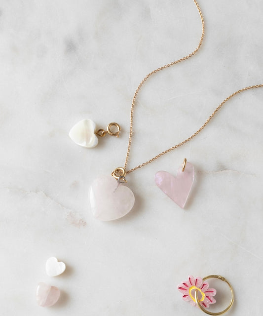 Heart-shaped jewelry, including pendants and charms, arranged on a white marble surface. A WALD World Rose quartz heart charm is showcased on a delicate gold chain, with additional pieces nearby. A light rose flower ring is also shown, enhancing the elegant display with its subtle beauty.