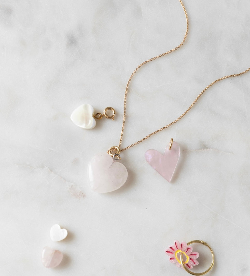 Heart-shaped jewelry, including pendants and charms, arranged on a white marble surface. A WALD World Rose quartz heart charm is showcased on a delicate gold chain, with additional pieces nearby. A light rose flower ring is also shown, enhancing the elegant display with its subtle beauty.