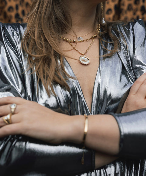 Wearing a metallic silver long-sleeve dress and layered WALD Berlin's handmade gold jewelry with pearl accents, including the Drop It Like It’s Hot Shell Necklace, arms crossed, against a leopard print background.