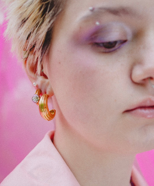 A person with short, light hair wears WALD Berlin’s SAMPLE SALE Pop Bi Color Crystal Earrings, a pink shirt, and purple eyeshadow, set against a pink background.