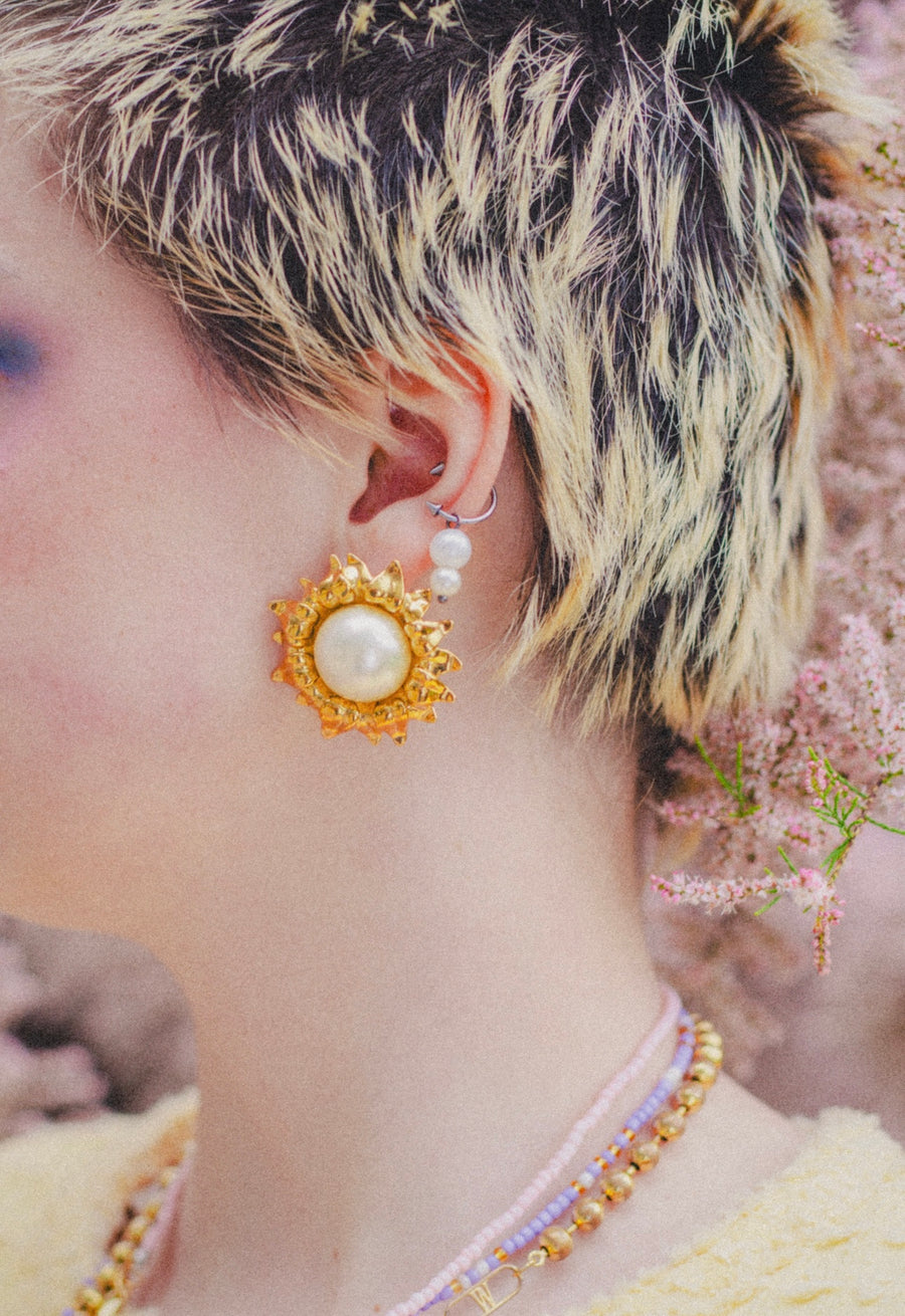 Close-up of a person with short, bleached and dark hair wearing WALD Berlin's SAMPLE SALE Pop Soleil Earrings—bold statement jewelry styled with a pearl ear cuff and layered beaded necklaces.
