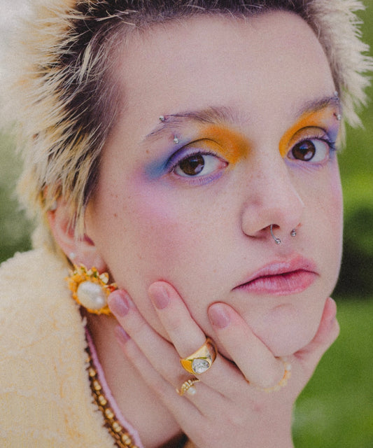 Person with short, spiky blond hair and colorful eye makeup rests their chin on their hand, wearing WALD Berlin's Mom Swarovski Heart Ring and gold jewelry, looking directly at the camera.