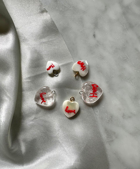 Five heart-shaped charms with "love" in red rest on gray marble beside shiny silver fabric, perfectly complementing the WALD Berlin Hi Mama pearl heart necklace.