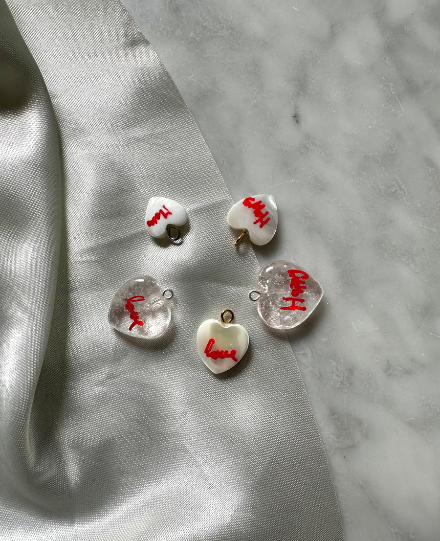 Five heart-shaped charms with "love" in red rest on gray marble beside shiny silver fabric, perfectly complementing the WALD Berlin Hi Mama pearl heart necklace.