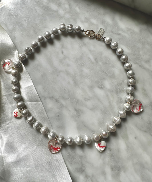 The WALD Berlin Hi Mama pearl heart necklace features five clear, red-lettered heart charms on gold-plated pearls—an elegant option for Mother's Day, beautifully displayed on marble.