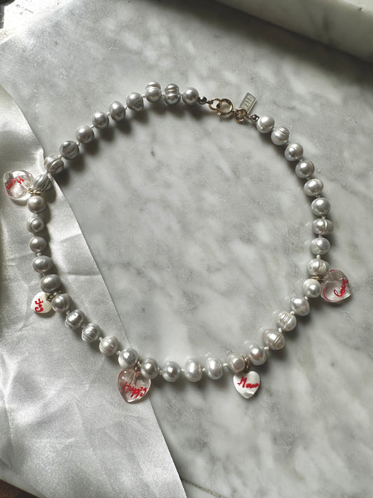 The WALD Berlin Hi Mama pearl heart necklace features five clear, red-lettered heart charms on gold-plated pearls—an elegant option for Mother's Day, beautifully displayed on marble.