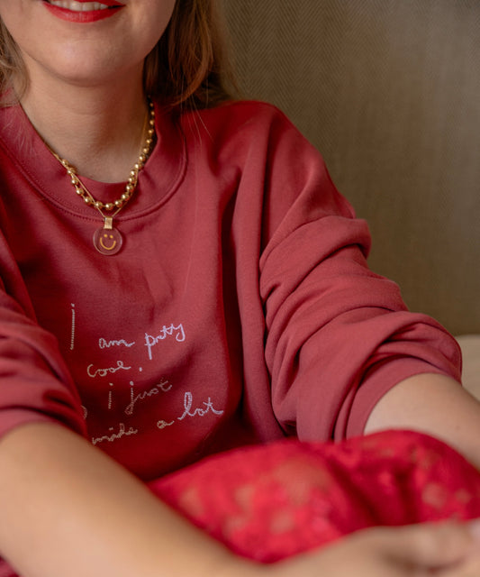 A person wearing a limited edition PRETTY COOL Jumper by WALD Berlin, paired with a beaded necklace and red lipstick, is seated with their arms crossed.
