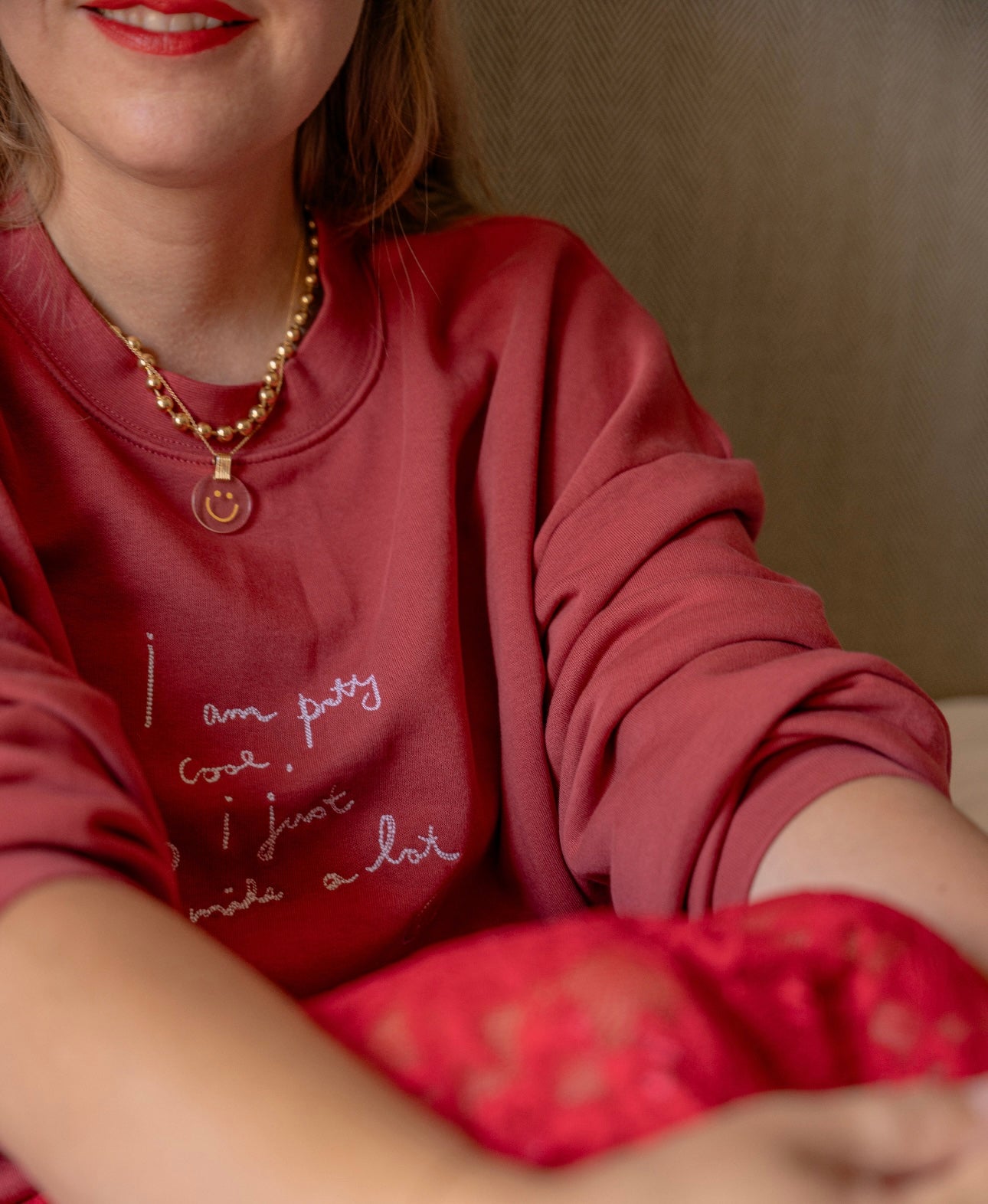 A person wearing a limited edition PRETTY COOL Jumper by WALD Berlin, paired with a beaded necklace and red lipstick, is seated with their arms crossed.