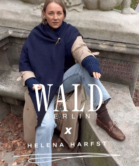 Eine Frau, die einen handgefertigten „Homeland“-Schal von WALD Berlin x Helena Harfst in Größe XL, einen Trenchcoat, blaue Jeans und braune Loafer trägt, sitzt auf einem Steinvorsprung in einem Park.