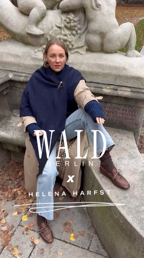 Eine Frau, die einen handgefertigten „Homeland“-Schal von WALD Berlin x Helena Harfst in Größe XL, einen Trenchcoat, blaue Jeans und braune Loafer trägt, sitzt auf einem Steinvorsprung in einem Park.