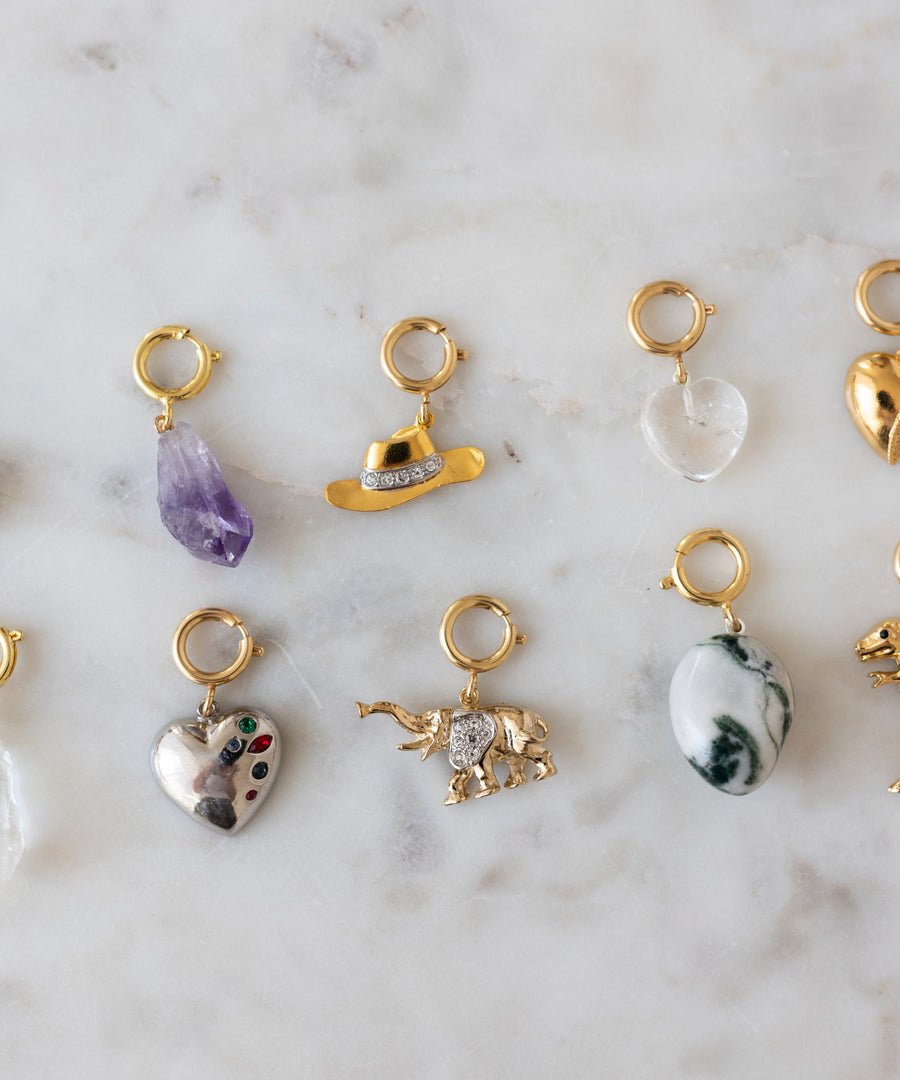Nine gold jewelry charms—featuring shapes like hearts, stones, a cowboy hat, and an elephant—are displayed on marble as the perfect inspiration for your next DIY Schmuck-Workshop x MINI im KaDeWe Berlin by WALD Berlin or your Charm DIY project.