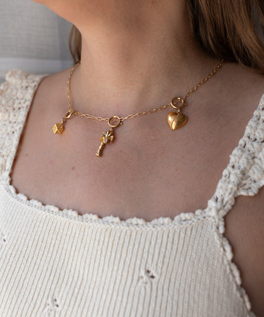Eine Goldkette von WALD Berlin mit drei Anhängern – einem Würfel, einem Schlüssel und einem Herz –, die über einem weißen, gehäkelten Tanktop getragen wird. Dieses Schmuckstück ist vom DIY-Schmuck-Workshop x MINI im KaDeWe Berlin inspiriert und verleiht jedem Look handgefertigte Eleganz.