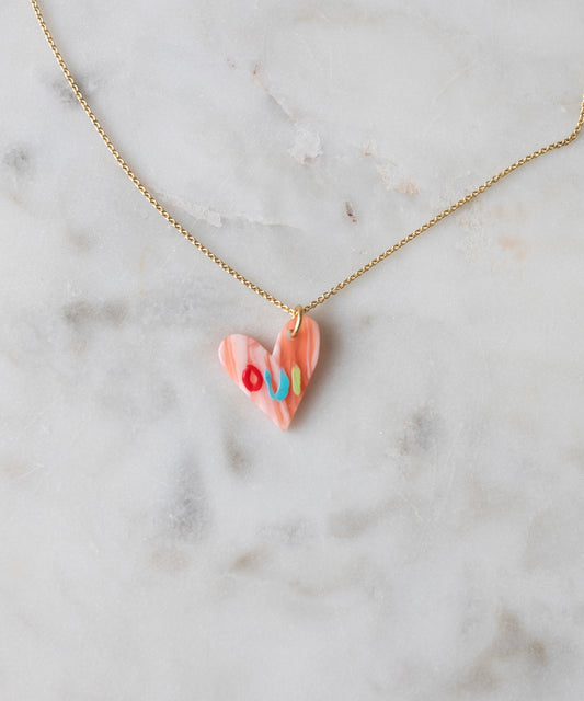 WALD Berlin's Pledges Of Tenderness MOM Necklace features a gold heart, pink marbled pendant, and colorful accents inspired by a handwritten love note, elegantly displayed on a light marble surface.