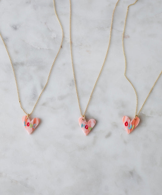 Three WALD Berlin "Pledges Of Tenderness MOM" gold heart necklaces with pink, heart-shaped pendants and colorful floral designs are displayed on white marble. These charming pieces are made in Germany.
