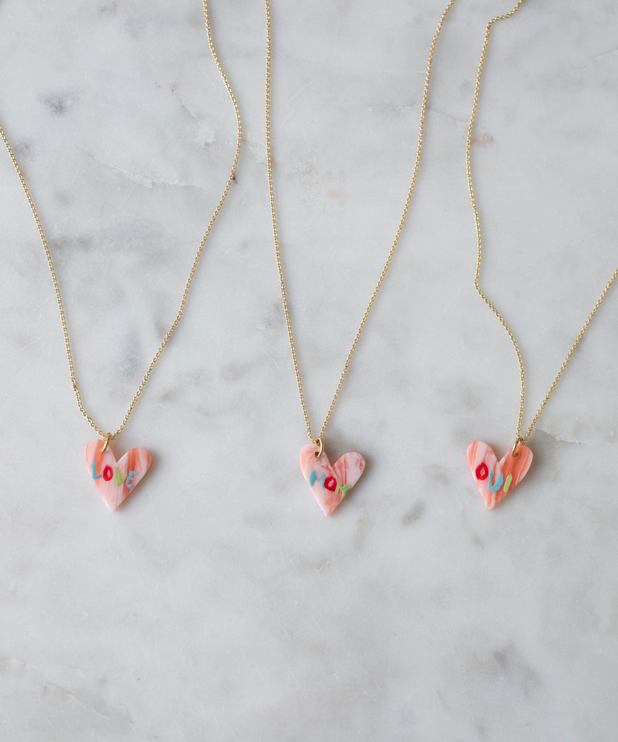 Three WALD Berlin "Pledges Of Tenderness MOM" gold heart necklaces with pink, heart-shaped pendants and colorful floral designs are displayed on white marble. These charming pieces are made in Germany.