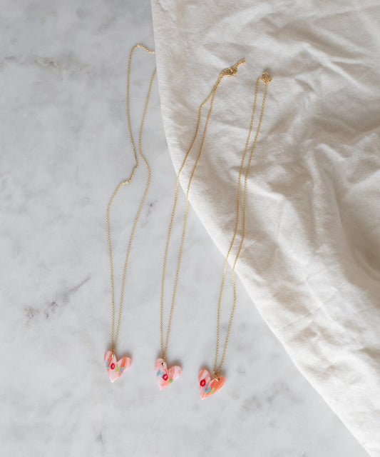 Three WALD Berlin Pledges Of Tenderness MOM gold heart necklaces with pink pendants are displayed on marble beside a white cloth. These elegant German-made pieces include a handwritten love note design.