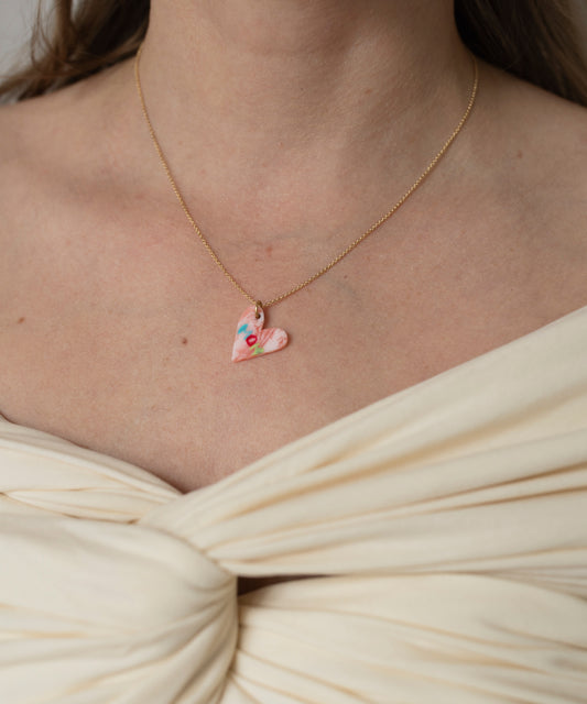 A close-up of a person wearing a cream off-the-shoulder top and the WALD Berlin Pledges Of Tenderness MOM Necklace, featuring a gold handwritten "love" note with a pink heart-shaped pendant.