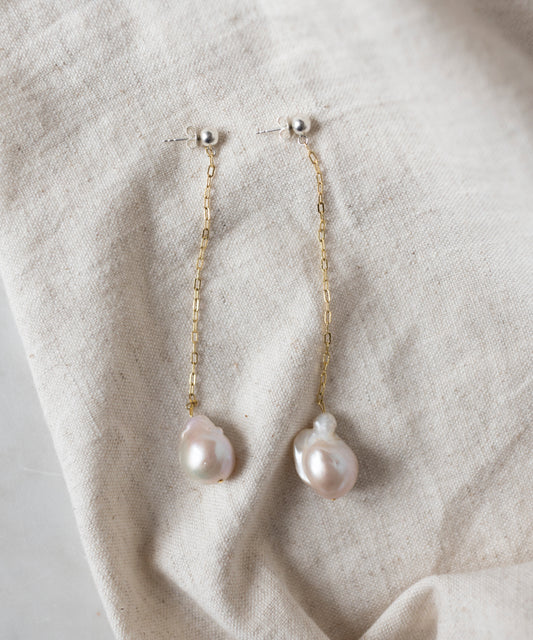 The WALD Berlin Happy Holidays Pearl Drop Earrings feature gold chains and irregular-shaped pearl pendants, shown on a beige fabric background.