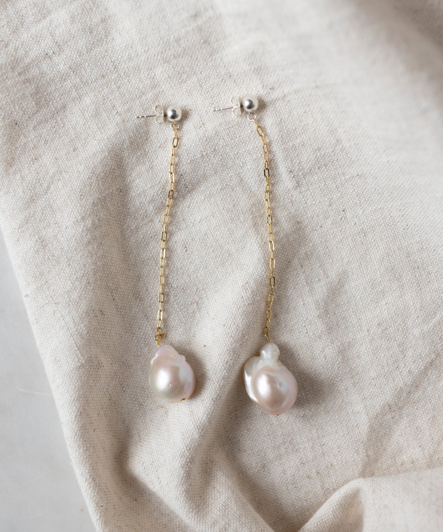 The WALD Berlin Happy Holidays Pearl Drop Earrings feature gold chains and irregular-shaped pearl pendants, shown on a beige fabric background.