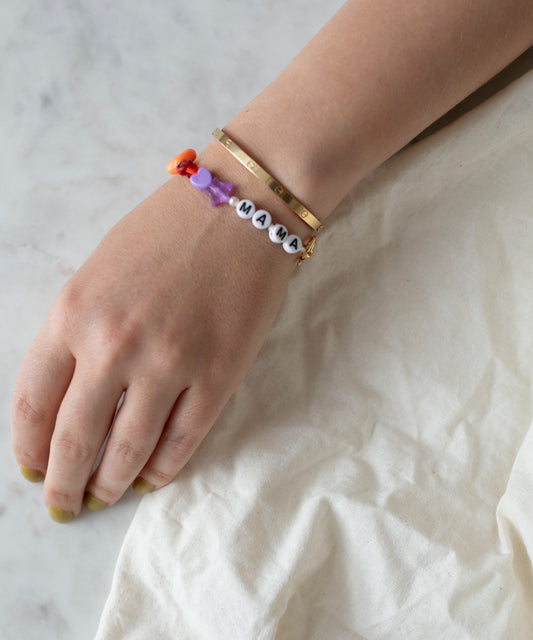 A hand rests on a white surface, adorned with a gold bangle and the WALD Berlin Personalized bracelet featuring colored shapes and white beads that spell “MAMA.”.
