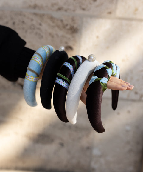 A hand displays WALD Berlin's SAMPLE SALE Baroque Pearl Black Headband—one of five stylish, padded headbands against a stone wall. Made in Germany, these accessories come in black, blue, white, striped designs, and more.