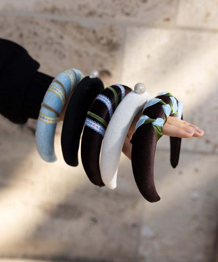 A hand displays WALD Berlin's SAMPLE SALE Baroque Pearl Black Headband—one of five stylish, padded headbands against a stone wall. Made in Germany, these accessories come in black, blue, white, striped designs, and more.