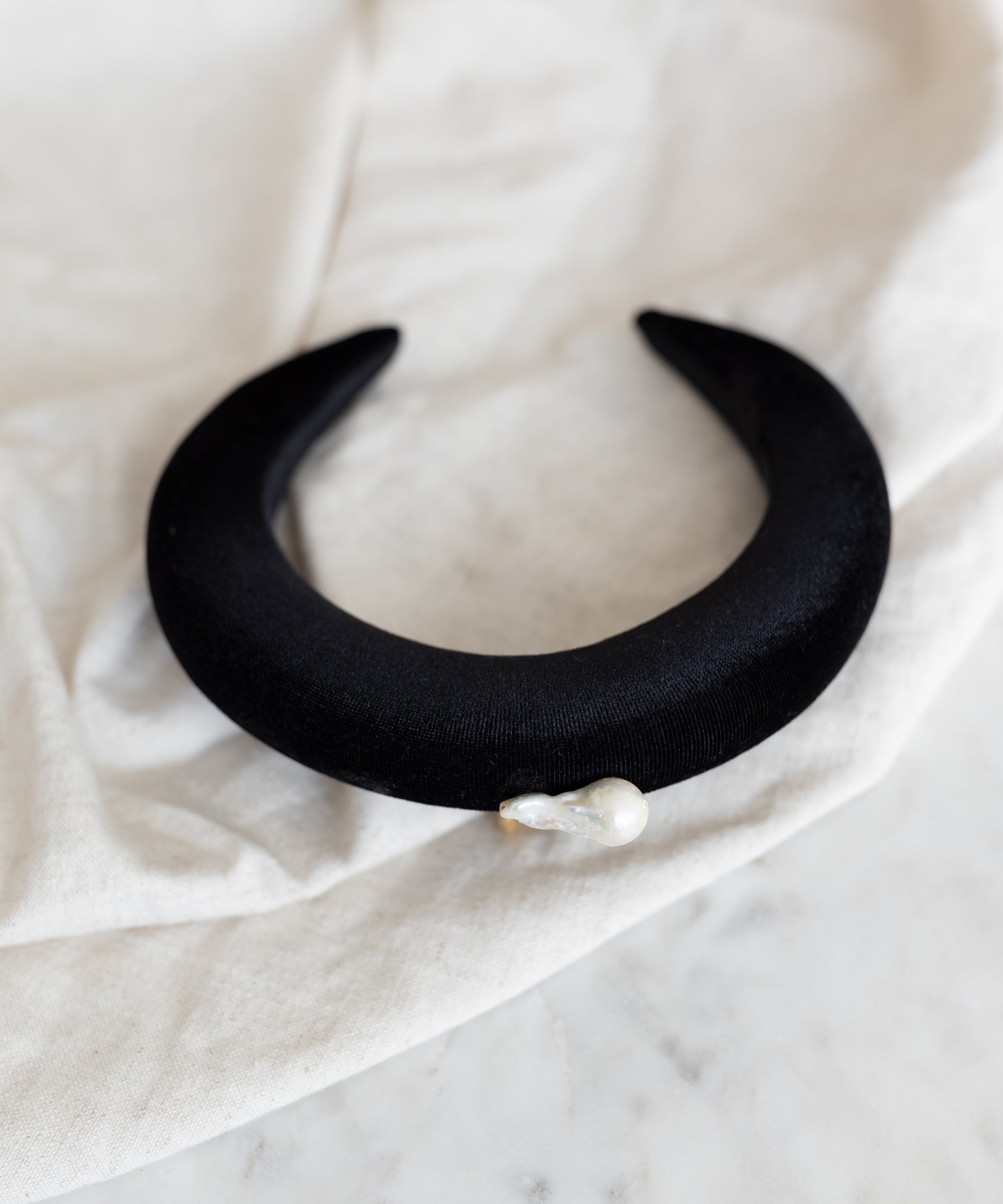 The WALD Berlin SAMPLE SALE Baroque Pearl Black Headband, featuring a black crescent shape with an irregular baroque pearl, is displayed on a light fabric surface.