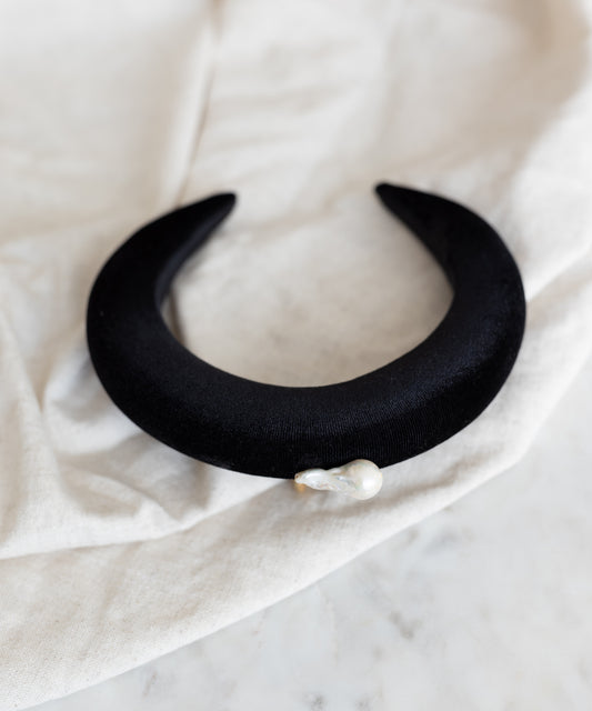 The WALD Berlin SAMPLE SALE Baroque Pearl Black Headband, featuring a black crescent shape with an irregular baroque pearl, is displayed on a light fabric surface.