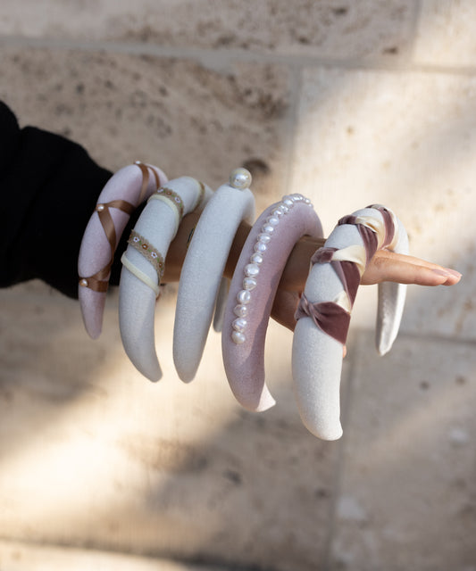 A hand displays the SAMPLE SALE Headband Knot White by WALD Berlin against a light stone background. This handmade-in-Germany piece features padded velvet in pastel tones, with pearl and ribbon details for a uniquely elegant look.
