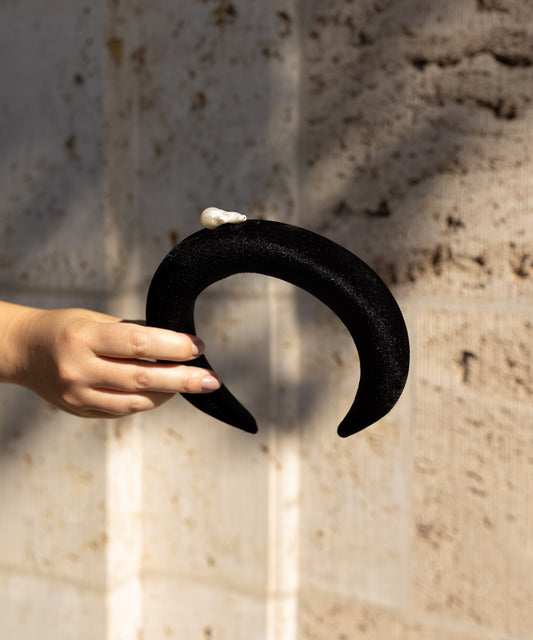 A hand holds the WALD Berlin SAMPLE SALE Baroque Pearl Black Headband, featuring a pearl-like detail, against a textured beige wall.