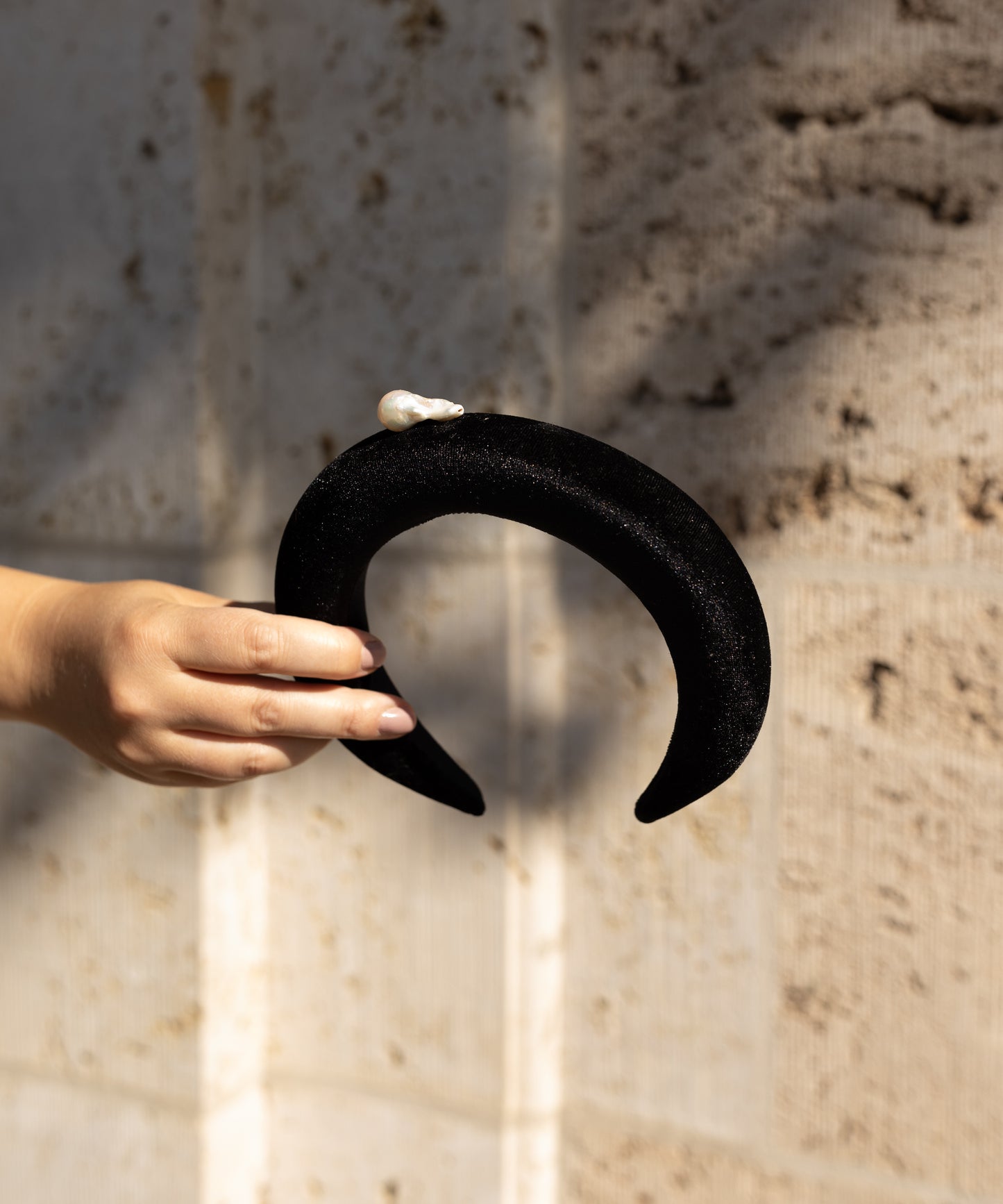A hand holds the WALD Berlin SAMPLE SALE Baroque Pearl Black Headband, featuring a pearl-like detail, against a textured beige wall.