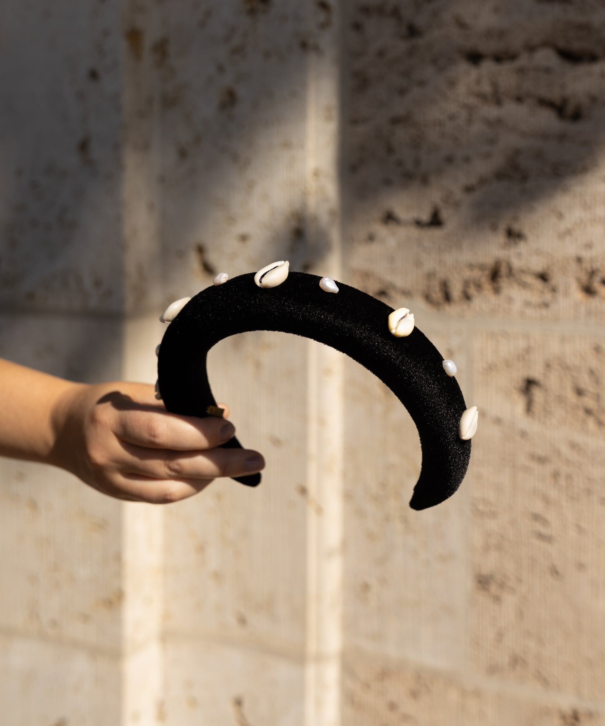 A hand holds the SAMPLE SALE Simone de Beauvoir Black Headband by WALD Berlin—black velvet with cowrie shells—set against a beige textured wall; a unique piece from our limited collection.