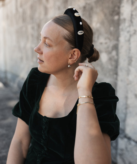 A woman wearing a black velvet dress and the SAMPLE SALE Simone de Beauvoir Black Headband by WALD Berlin, sitting against a textured concrete wall and looking to the side.