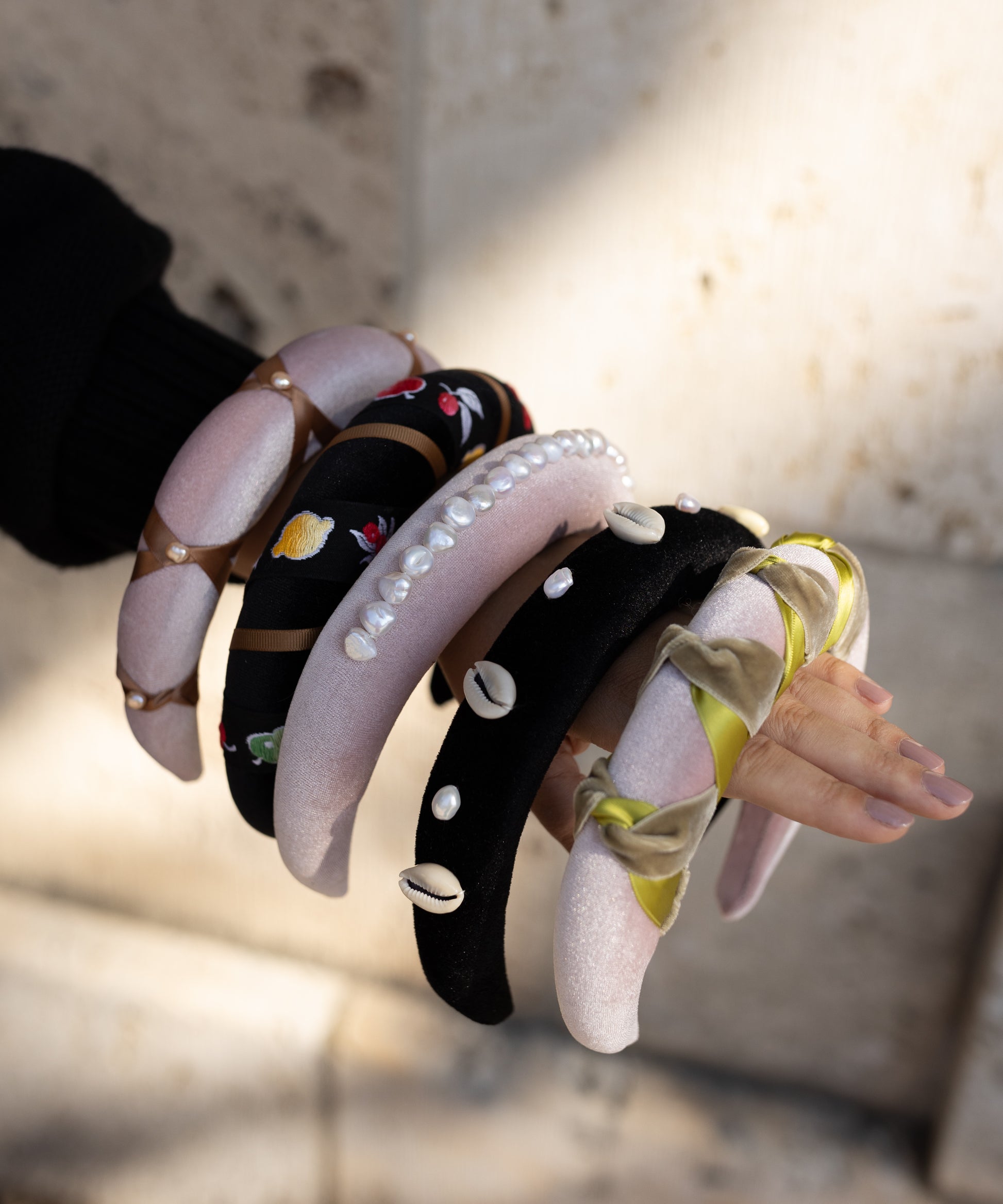 A person’s arm shows off several padded WALD Berlin headbands, like the SAMPLE SALE Simone de Beauvoir Black Headband, in unique designs—pearls, embroidery, shells, and ribbons—against a blurred stone background.