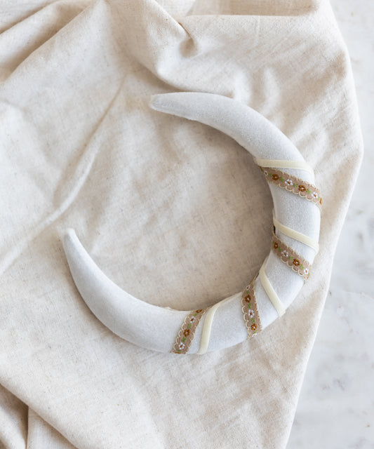 The SAMPLE SALE Headband Swirl White by WALD Berlin, featuring a crescent shape with sequin and ribbon details in white velvet, is displayed on a light beige fabric background.