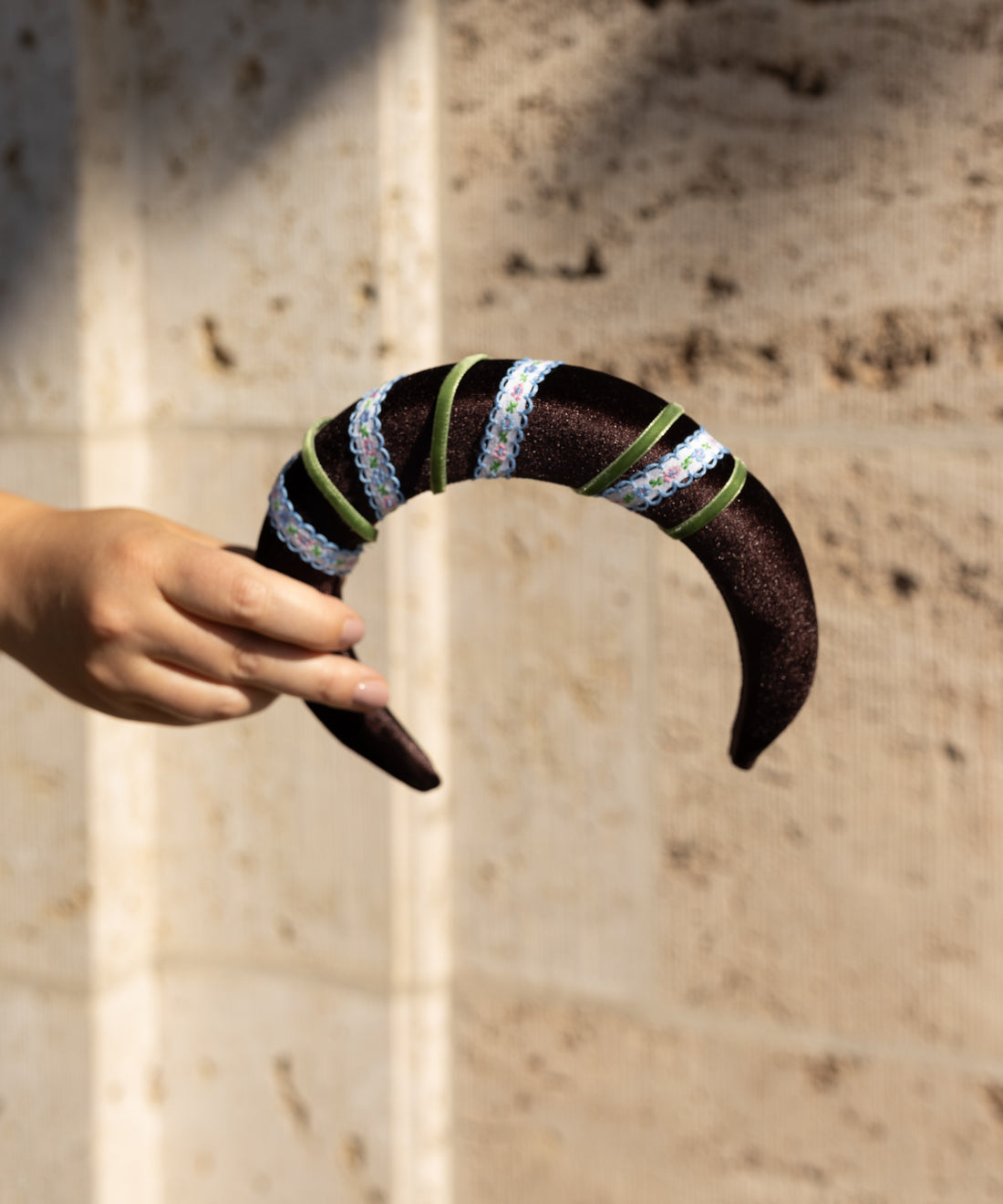 A hand holds the WALD Berlin SAMPLE SALE Headband Swirl Dark Brown, a curved accessory adorned with green ribbon and iridescent sequins, set against a beige textured wall.