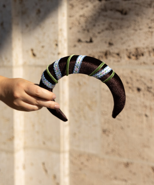 A hand holds the WALD Berlin SAMPLE SALE Headband Swirl Dark Brown, a curved accessory adorned with green ribbon and iridescent sequins, set against a beige textured wall.