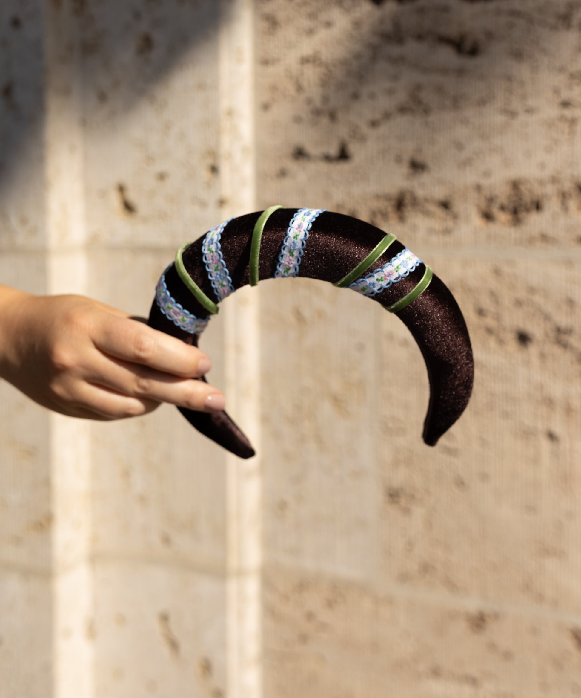 A hand holds the WALD Berlin SAMPLE SALE Headband Swirl Dark Brown, a curved accessory adorned with green ribbon and iridescent sequins, set against a beige textured wall.
