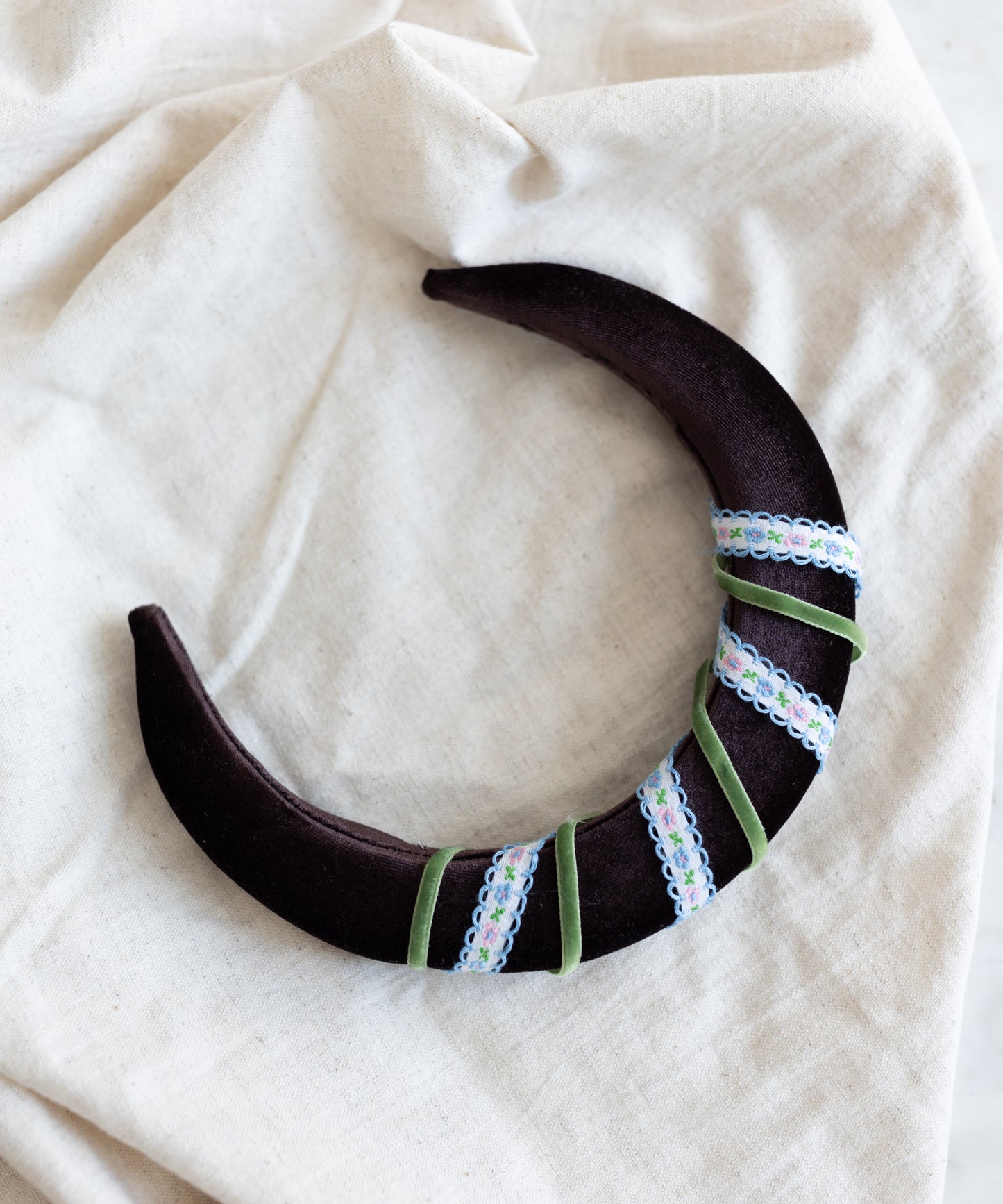The SAMPLE SALE Headband Swirl Dark Brown by WALD Berlin, handmade in Germany, features a dark crescent shape with green ribbon and lace, displayed on a cream-colored fabric background.