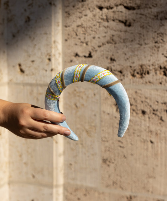A hand holds the WALD Berlin SAMPLE SALE Headband Swirl Light Blue, a crescent-shaped velvet piece with embroidered designs, shown against a textured stone wall background.
