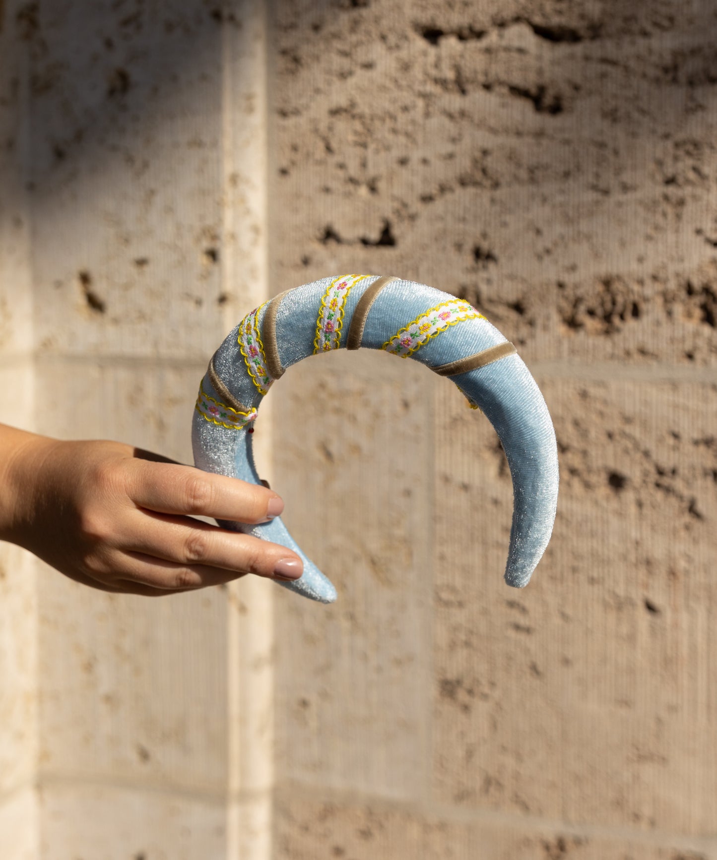 A hand holds the WALD Berlin SAMPLE SALE Headband Swirl Light Blue, a crescent-shaped velvet piece with embroidered designs, shown against a textured stone wall background.