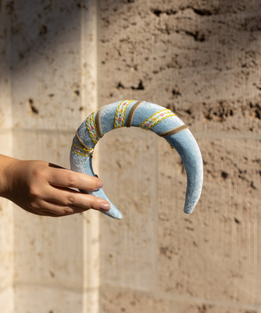 A hand holds the WALD Berlin SAMPLE SALE Headband Swirl Light Blue, a crescent-shaped velvet piece with embroidered designs, shown against a textured stone wall background.
