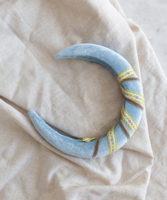The WALD Berlin SAMPLE SALE Headband Swirl Light Blue features a soft, crescent shape in blue velvet with yellow and pink ribbon accents, displayed on a light beige fabric background.