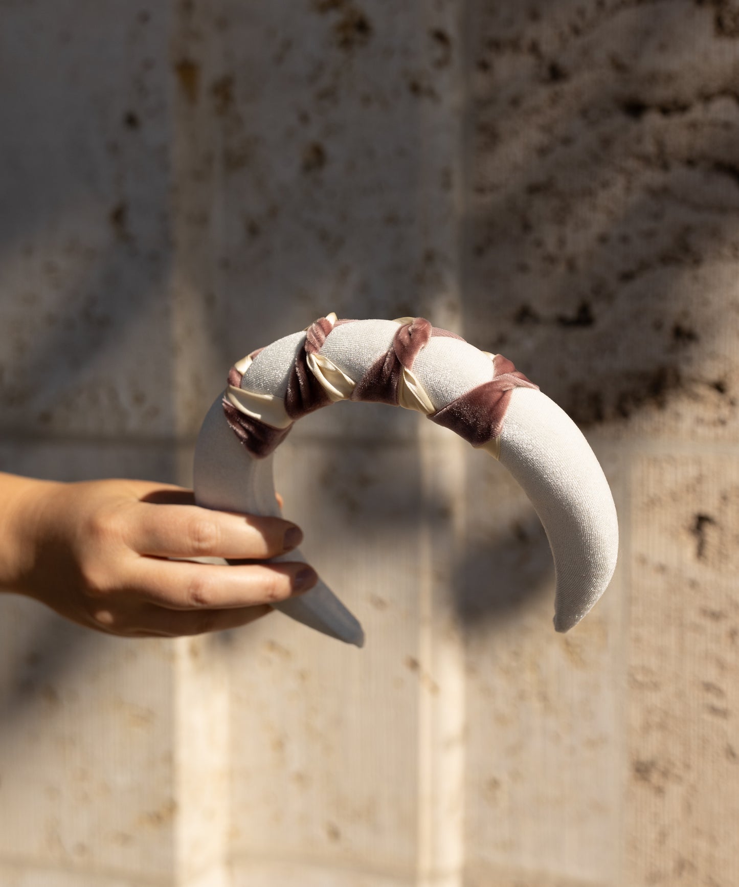 A hand holds the WALD Berlin SAMPLE SALE Headband Knot White, a crescent-shaped velvet piece with maroon ribbon and gold accents, set against a textured beige wall.