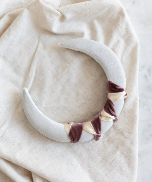The SAMPLE SALE Headband Knot White by WALD Berlin, a handmade padded crescent-shaped piece, features twisted beige and mauve velvet accents on light gray fabric and is displayed on cream-colored cloth.