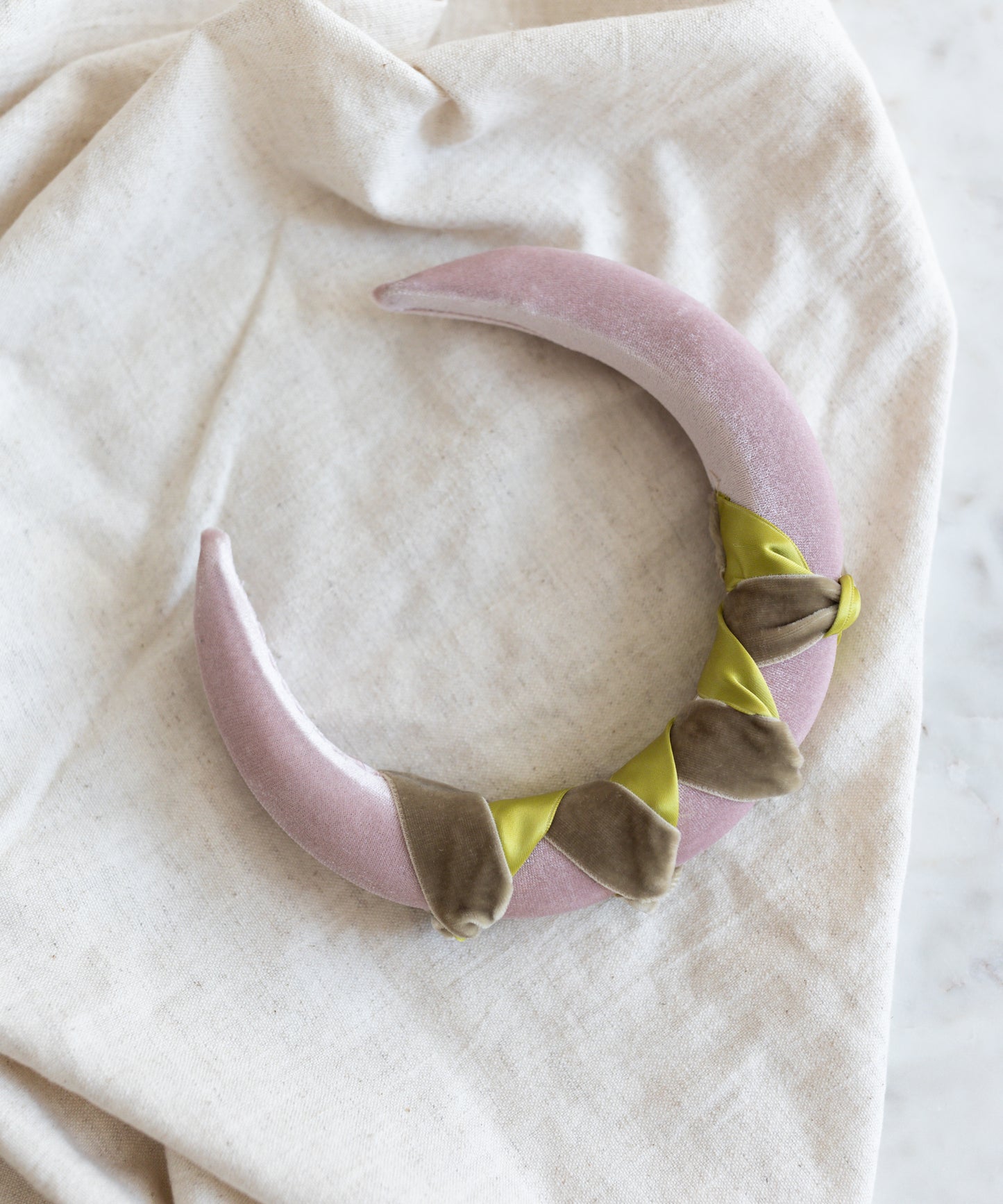 The SAMPLE SALE Headband Knot Rose by WALD Berlin, a crescent-shaped, pale pink plush accessory with green and brown accents, handmade in Germany, rests on a beige cloth.