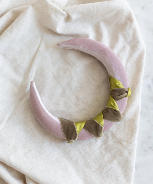 The SAMPLE SALE Headband Knot Rose by WALD Berlin, a crescent-shaped, pale pink plush accessory with green and brown accents, handmade in Germany, rests on a beige cloth.