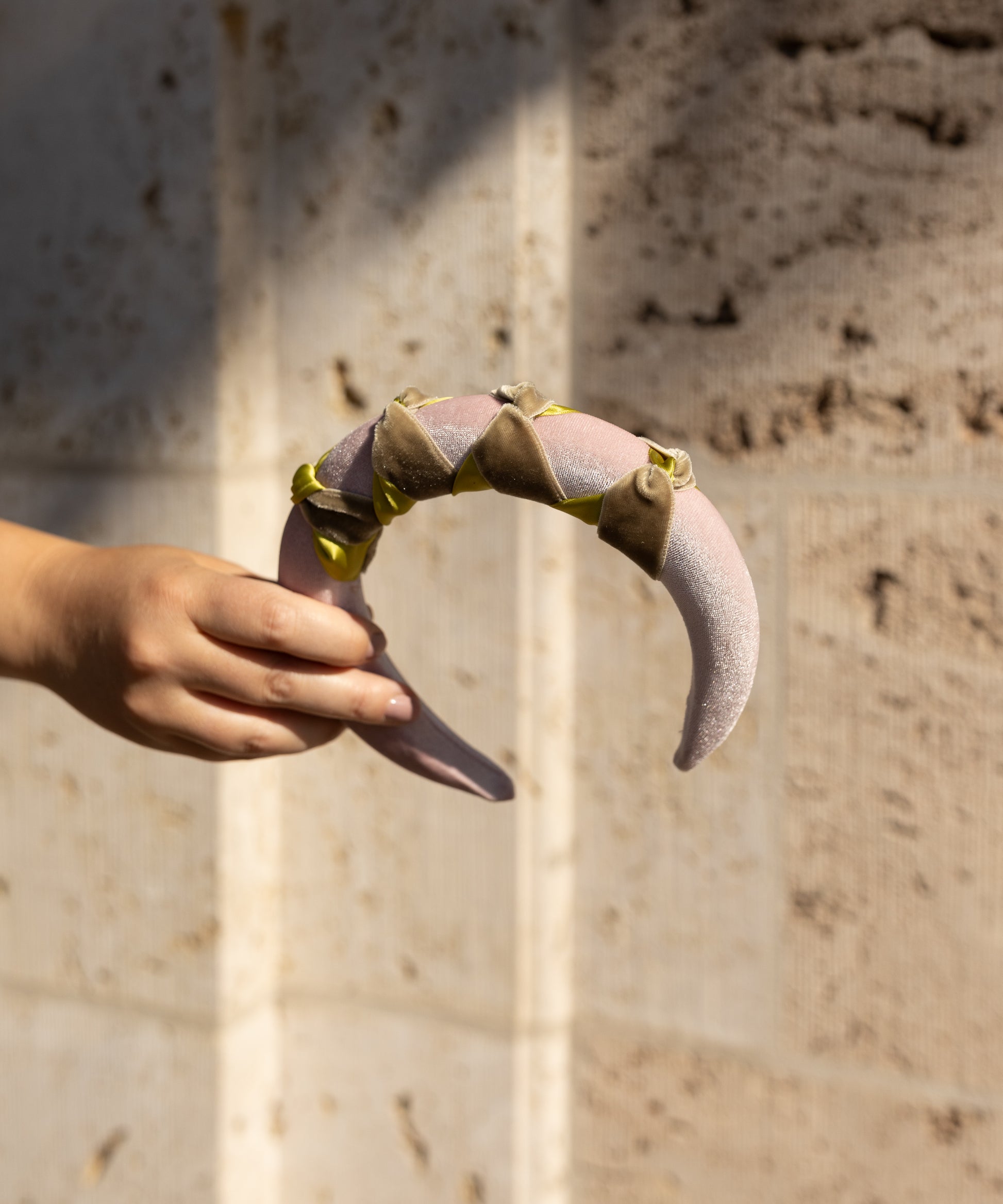A hand displays the SAMPLE SALE Headband Knot Rose by WALD Berlin, a crescent-shaped pink accessory with green accents and triangular details, set against a textured beige wall.